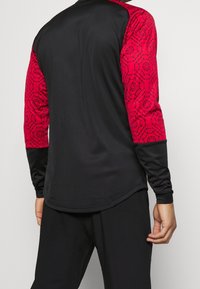 Long-sleeve athletic shirt featuring a black body with red patterned sleeves, smooth texture, and contrasting black cuffs.