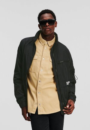 Giacca nera con zip e polsini elasticizzati sopra una camicia beige con colletto. La camicia presenta due tasche sul petto e bottoni a pressione. I jeans scuri completano il look.