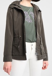 Parka en kaki avec une capuche à cordon de serrage, fermeture éclair et deux poches à l'avant. Portée par-dessus un t-shirt graphique vert et un pantalon blanc.