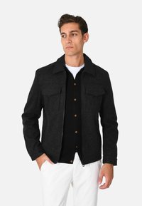 Giacca nera in misto lana con colletto, due tasche sul petto e chiusura con zip. Indossata sopra una camicia nera con bottoni e pantaloni bianchi.