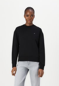 Felpa nera con collo a giro e polsini a coste. Presenta un piccolo logo bianco sul petto. Indossata con jeans grigi chiari.