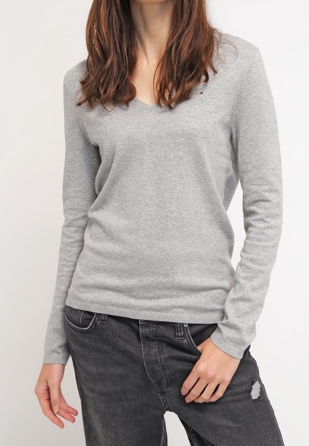 Pull gris à col en V en matière douce, avec manches longues et silhouette ajustée, associé à un jean gris foncé déchiré.