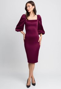 Bordowa dopasowana sukienka z kwadratowym dekoltem, bufiastymi rękawami i długością do kolan. Gładki materiał i sylwetka podkreślająca figurę. Czarne szpilki.