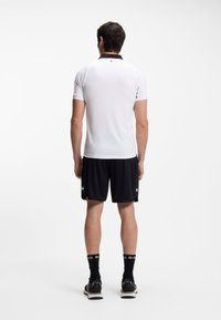 Weißes Poloshirt mit kurzen Ärmeln und schwarzem Kragen, kombiniert mit schwarzen Shorts. Das Outfit wird durch schwarze Socken und sportliche Schuhe ergänzt.