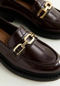 Brązowe błyszczące skórzane mokasyny z wyraźnym złotym łańcuchem na pasku i teksturowaną czarną gumową podeszwą.