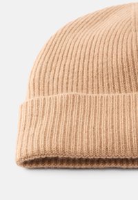 Beige knit beanie hat made of soft, ribbed material. Features a rolled brim and a textured surface with visible stitching details.