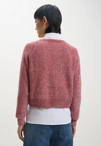 Maglione rosa testurizzato e rigato con un taglio corto e maniche raglan, indossato sopra una camicia bianca. Il motivo a maglia è ben visibile.