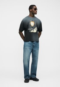 Άνδρας που φοράει μαύρο t-shirt με σχέδιο, μπλε τζιν, μαύρα αθλητικά παπούτσια, γυαλιά ηλίου και αλυσίδα στο λαιμό, στέκεται με τα χέρια πίσω από την πλάτη.