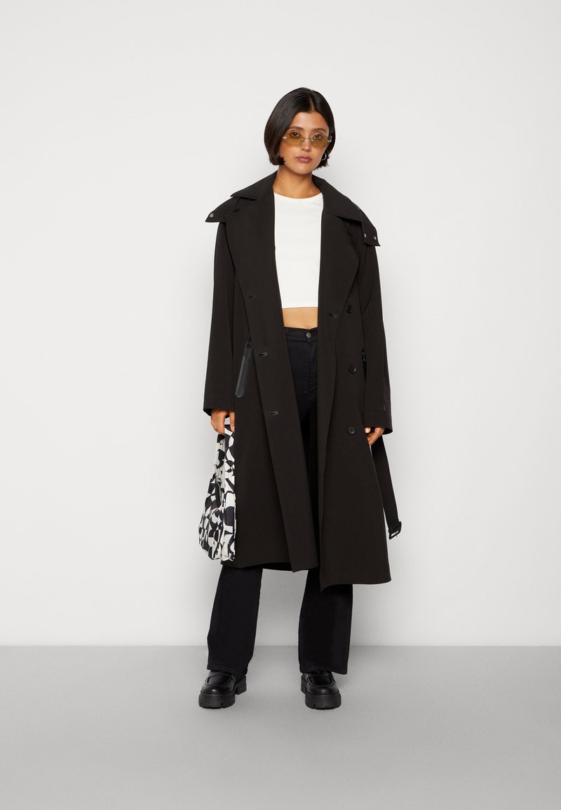 Cappotto trench nero con vita cintata, ampi risvolti e chiusura doppiopetto. Indossato sopra una maglietta bianca a maniche corte, abbinato a pantaloni neri e una borsa con motivo.