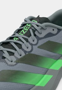 Γκρι αθλητικό παπούτσι με πράσινες λεπτομέρειες, που διαθέτει υφασμάτινο επάνω μέρος, κορδόνια ίδιου χρώματος και τρεις χαρακτηριστικές σκούρες πράσινες ρίγες στο πλάι.