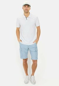 Hvid polo T-shirt med sorte knapdetaljer, lyseblå cargo-shorts og hvide sneakers. Modellen bærer en lyseblå kasket.