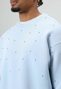 Sweat-shirt bleu clair avec un col rond, une coupe ample et des points noirs éparpillés pour la texture et les détails. Tissu doux avec poignets côtelés.