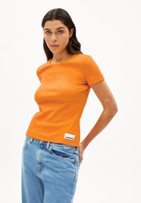 Oranje geribde t-shirt met een aansluitend ontwerp en korte mouwen. Bevat een wit label aan de onderkant links. Gecombineerd met lichtblauwe jeans.