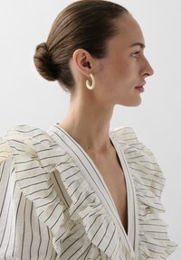 Blusa crema con volant e righe navy, scollatura a V profonda, abbinata a grandi orecchini a cerchio giallo pallido, che evidenziano il design curvo e la texture liscia.