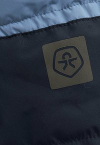 Dunkelblauem Stoff mit glatter Textur zeigt ein hellblaues Paneel oben und einen schwarzen Logopatch mit dem Symbol einer menschlichen Figur.