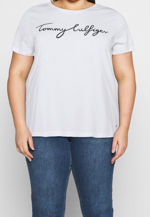 Person wearing a white short-sleeve shirt with "Tommy Hilfiger" script logo and blue denim jeans, shown from neck to hips.