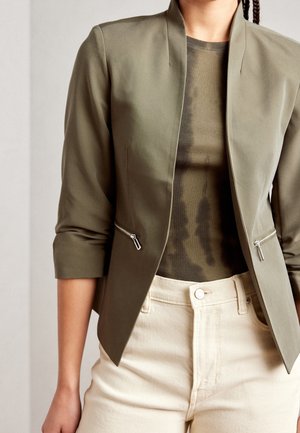 Mujer con blazer corta de color verde oliva con bolsillos con cremallera sobre top de canalé en tono oliva y pantalones de talle alto color blanco roto.