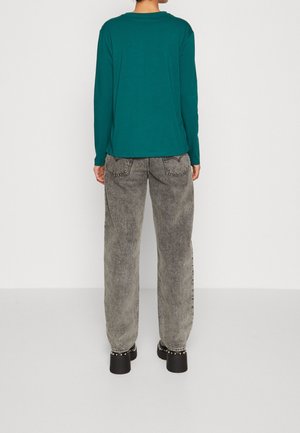 Henkilö, jolla on päällään teekkarinvihreä pitkähihaisen paita, väljät harmaat happopesulahkeiset farkut ja mustat korokepohjaiset kengät hopeisilla niiteillä, seisoo valkoista taustaa vasten.