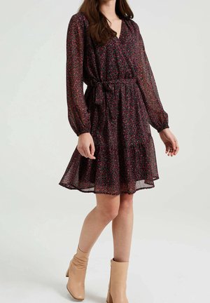 Vrouw die een donker bloemenpatroon jurk met lange mouwen, een gestrikte taille en beige enkellaarzen draagt, staat tegen een effen achtergrond.