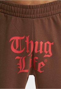 Brūni sporta bikses ar elastīgu jostasvieta un lielu sarkanu "Thug Life" grafiku stilizētā fontā priekšpusē, izgatavotas no mīkstas auduma.