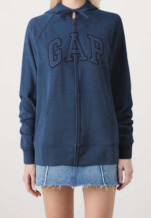 Osoba ubrana w niebieską bluzę z kapturem GAP na zamek błyskawiczny z troczkami oraz niebieską postrzępioną mini spódnicę jeansową, stojąca na jednolitym tle.