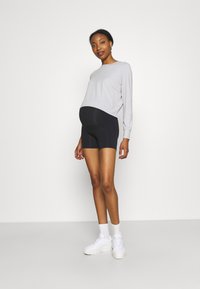 Un short de maternité noir et un haut à manches longues gris clair associés à des baskets blanches, se tenant devant un fond blanc uni.