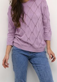 Kvinna med en lavendelfärgad stickad tröja med diamantformade utskärningar och ljusblå jeans, stående mot en enfärgad ljus vägg.