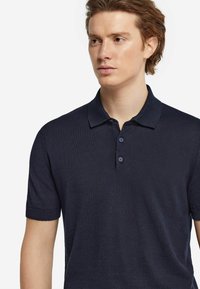 Marinblå polo t-shirt med mjuk textur, korta ärmar, krage och två knappar fram. Enkel design med uppvikt ärm.