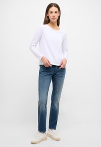 Weißes Langarmshirt mit Rundhalsausschnitt, kombiniert mit blauen Jeans und weißen Sneakers. Der Stoff wirkt glatt und lässig.