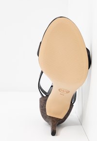 Vue de la semelle d'une chaussure à talon haut marron à motifs avec une semelle beige texturée et un talon pointu reposant contre une surface blanche.