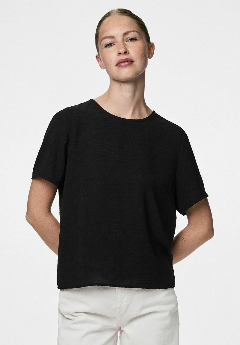 Top nero a maniche corte con vestibilità rilassata e scollatura rotonda, realizzato in un tessuto testurizzato. Indossato con pantaloni di colore chiaro.