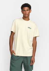 Crème-kleurige katoenen t-shirt met een teal golflogo en oranje tekst op de borst, gecombineerd met groene korte broeken.