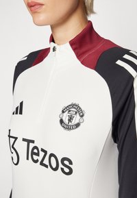 Fehér sportos zipzáras felső, amely bordó és fekete színblokkokkal rendelkezik. A Manchester United logója és a "Tezos" felirat fekete színben látható.