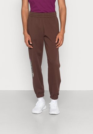 Pantalones de chándal marrones con cintura elástica y puños, que presentan un estampado gráfico en la pierna izquierda. Combinados con zapatillas deportivas blancas.