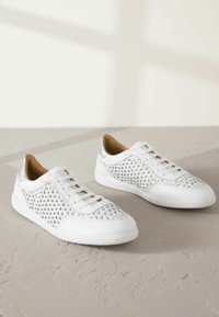 Weiße Sneakers mit gewebter Textur an den Seiten, metallischen silbernen Akzenten, flachen Schnürsenkeln und einer Gummisohle, auf einer neutralen Fläche platziert.