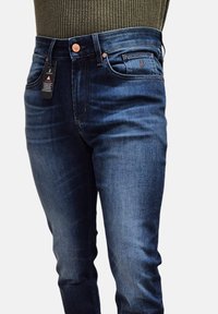 Jeans in denim blu scuro con vestibilità dritta, hardware in ottone e un'etichetta di stoffa attaccata alla vita. Dettagli di leggero sbiadimento.