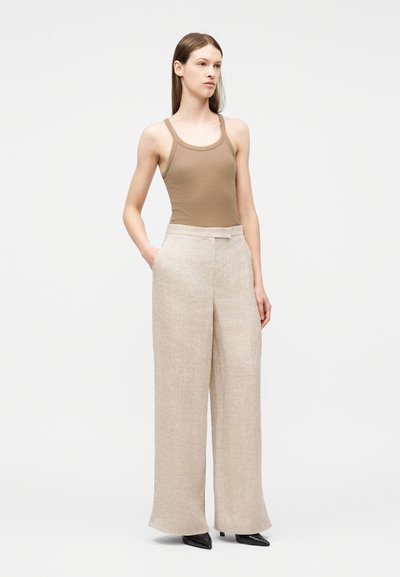 Femme debout les mains dans les poches, portant un haut sans manches beige, un pantalon large crème, et des chaussures noires pointues, sur un fond blanc uni.