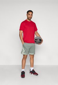 Κόκκινο κοντομάνικο μπλουζάκι με μαύρο λογότυπο, πράσινα υφασμάτινα σορτς με λευκές λεπτομέρειες, αθλητικά παπούτσια σε μαύρο και κόκκινο χρώμα και ένα μπάσκετ.