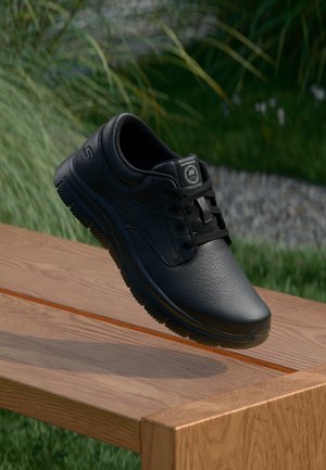 Zwarte leren Skechers sneaker met veters zwevend boven een houten bank buiten met gras- en kiezelachtergrond.