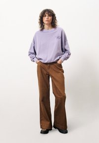 Lila übergroßer Pullover mit floralem Design auf der linken Brust, kombiniert mit weiten braunen Cordhosen. Schwarze Schuhe runden das Outfit ab.