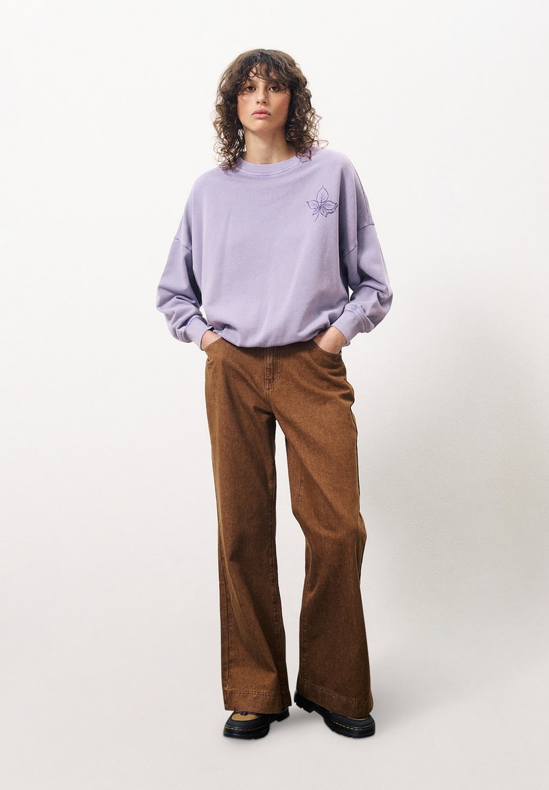 Lila übergroßer Pullover mit floralem Design auf der linken Brust, kombiniert mit weiten braunen Cordhosen. Schwarze Schuhe runden das Outfit ab.