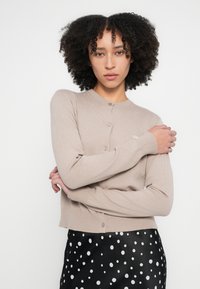 Bēšas krāsas adīts kardigans ar apaļu kakla izgriezumu un pogu aizdari. Tekstūra ir gluda, papildināta ar melnu svārku ar baltām pūtītēm.