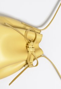 Gelbe Leder-Eimer-Tasche mit weicher Textur. Enthält dünne verstellbare Träger und ein Knotendetail an der Öffnung. Glatte Oberfläche.
