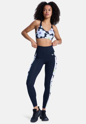 Brassière de sport fleurie noire avec des fleurs blanches et roses ; leggings taille haute bleu marine avec des bandes latérales fleuries ; tissu extensible et respirant.