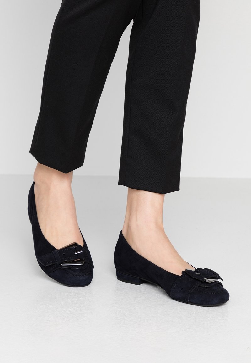 Pieds portant des mocassins noirs en daim à talon bas avec un détail de boucle argentée, associés à un pantalon noir court sur un sol clair.