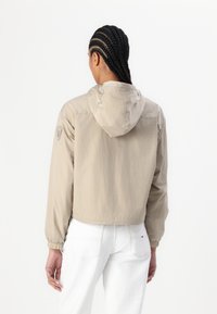 Jachetă bej scurtă cu glugă, mansete elastice și un patch cu logo pe brațul stâng. Materialul pare ușor și ușor texturat.