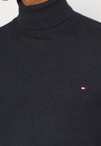 Pull à col roulé bleu marine foncé avec un petit logo rectangulaire rouge, blanc et bleu sur la partie gauche de la poitrine.