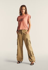 T-shirt rosa chiaro a maniche corte con una stampa grafica, abbinato a pantaloni cargo khaki larghi con molte tasche e polsini elastici.