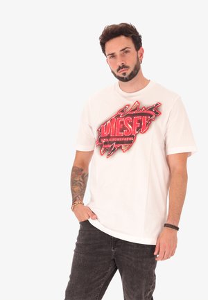 Homme avec barbe et bras tatoué portant un t-shirt Diesel blanc avec un graphique rouge et un jean noir, posant devant un fond blanc uni.
