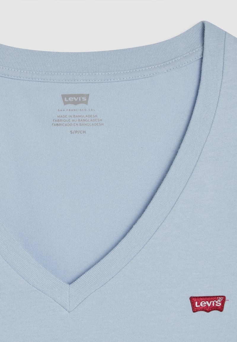 Camiseta de algodón azul claro con cuello en V y parche del logo rojo de Levis, con una textura suave y un corte estándar. Detalles de la etiqueta impresos en el interior del cuello.